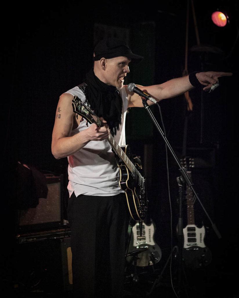 Kuvassa artisti SPK Toivonen lavalla, soittamassa kitaraa ja laulamassa, osoittaen yleisöä