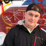 Mies seisoo graffitimaalatun seinän edessä ja hymyilee kameralle.