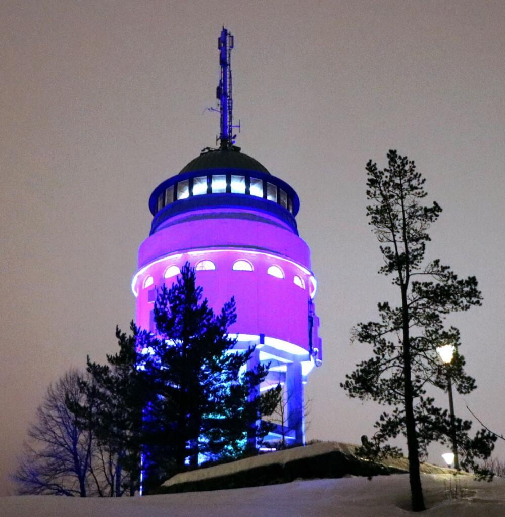 liilaksi valaistu hehkuva näköalatorni harmaata taivasta vasten, mustio puiden profiileja