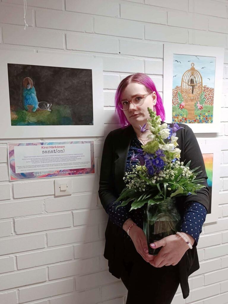 Taiteilijalla on sylissään kukkakimppu. Hän seisoo teostensa edessä näyttelyssään.